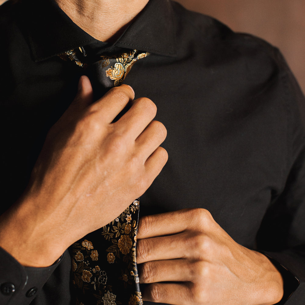 Person adjusting a black and gold tie with floral embroidery on a blurred background