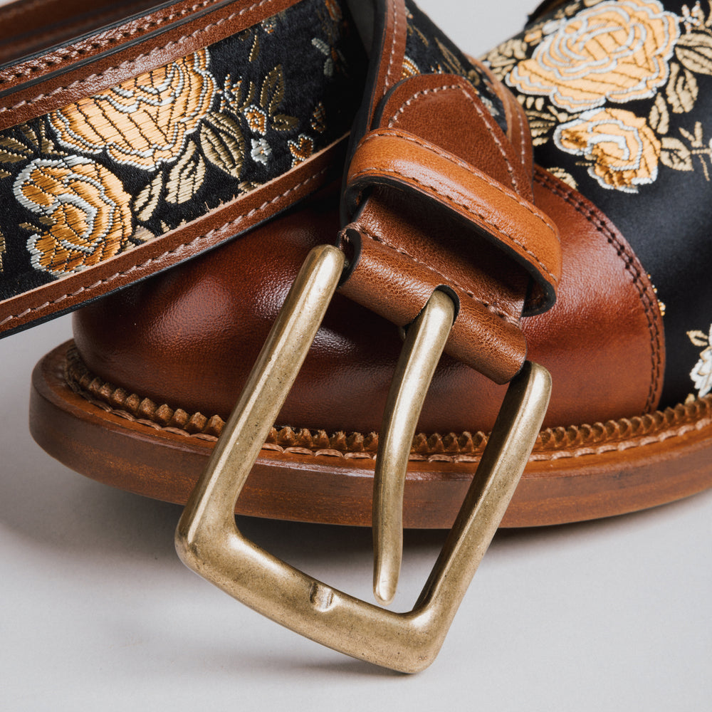 Brown leather belt with floral patterned top and brass buckle on a light gray background