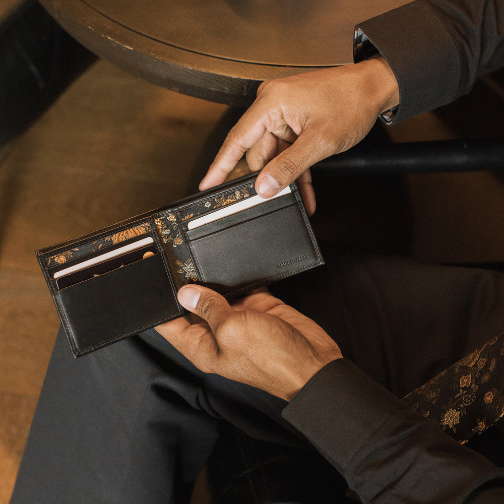 Person holding a black leather wallet with floral patterns, sitting on a wooden chair. - lifestyle